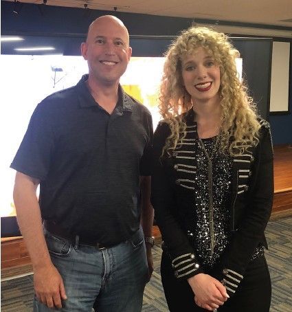 Man and woman posing. The man has a shaved head, the woman has curly blonde hair. They stand in front of a screen.