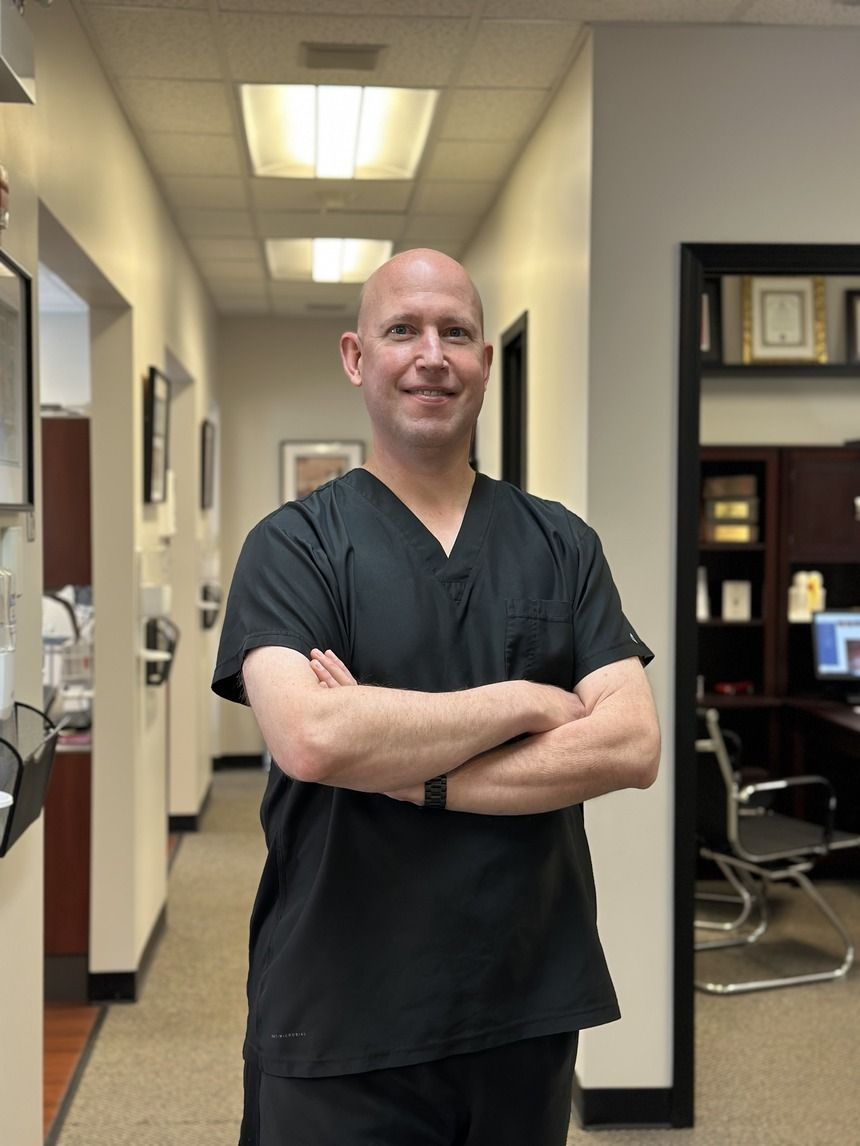 A man with a shaved head in black scrubs stands in a hallway with arms crossed. He's smiling and looking at the camera.