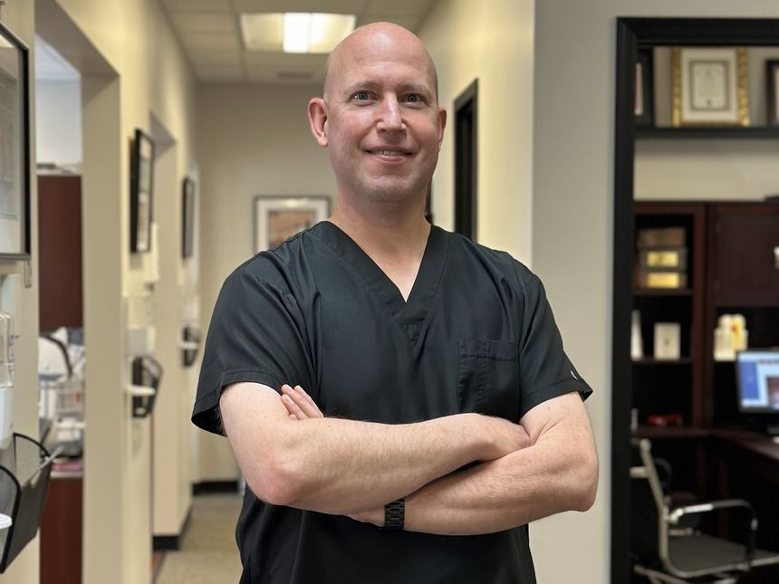 A smiling man in black scrubs stands in a hallway with arms crossed. He has short hair and is looking at the camera.