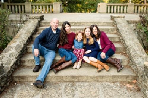 Family of five sits on stone steps, smiling at the camera. Man in blue shirt, women in burgundy and purple, and girls in dresses.