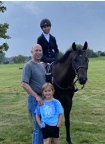 A family poses with a horse in a grassy field. A child in riding gear sits on the horse, while a man and another child stand below.