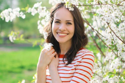 Woman with brown hair and a big smile in front of blossoming white flowers, wearing a red-and-white striped shirt.