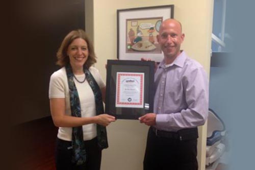 Woman and man holding framed certificate, smiling, in office setting. A cartoon is visible on the wall.