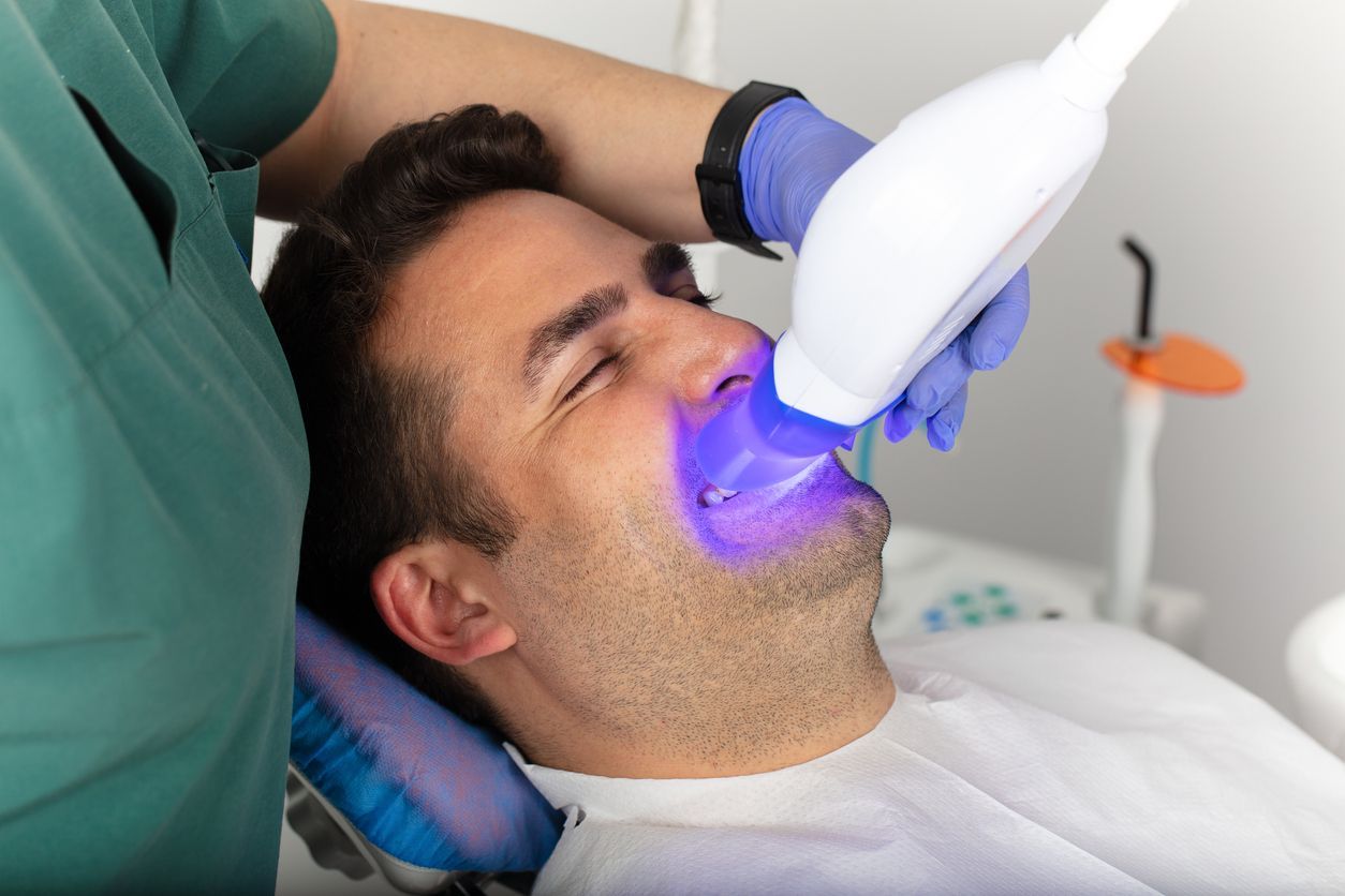 Man receiving teeth whitening treatment in a dental clinic. A dentist holds a blue light device to his teeth while he reclines with his eyes closed.