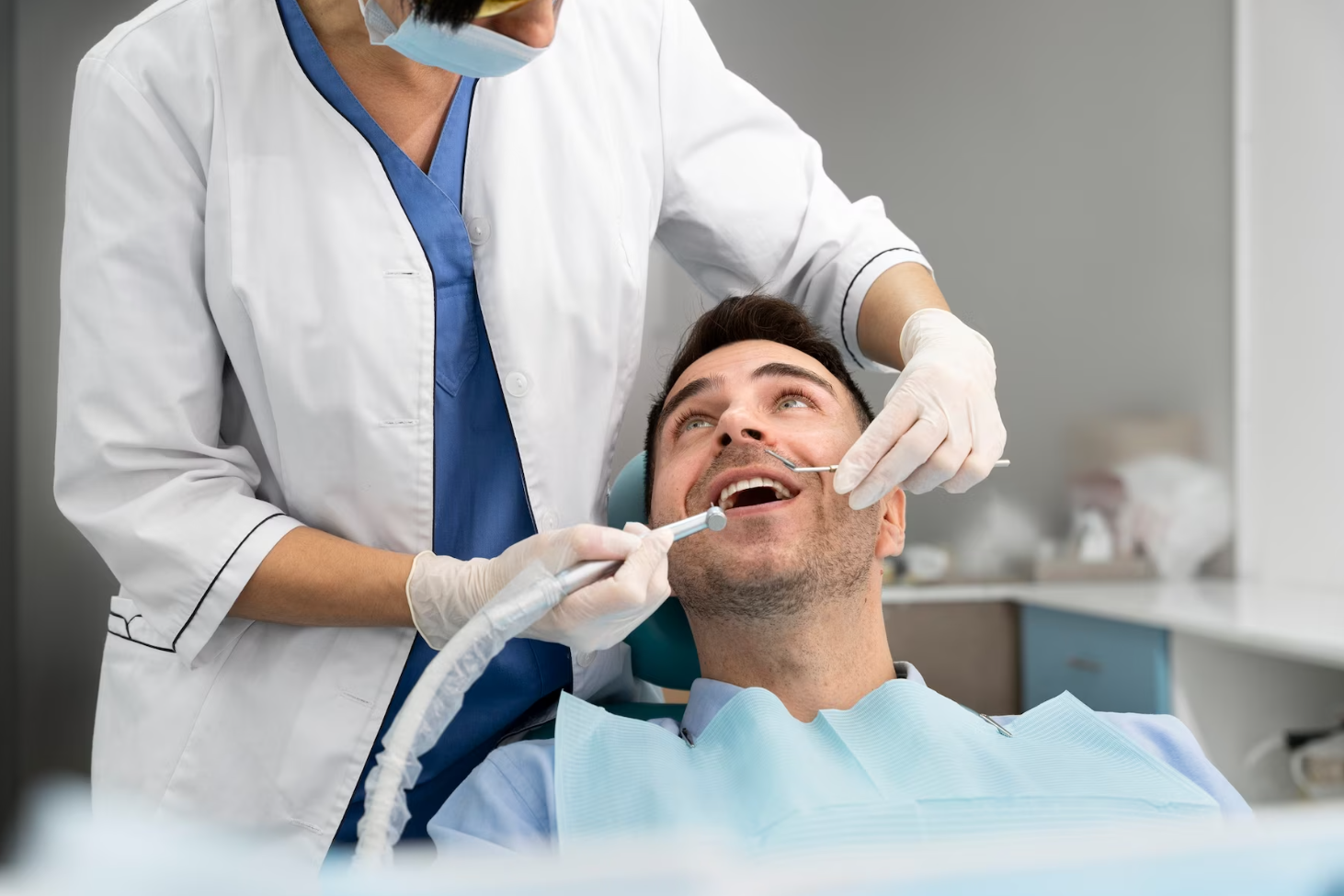 A dentist in a white coat and mask examines a patient's teeth, holding dental tools in a clinic.