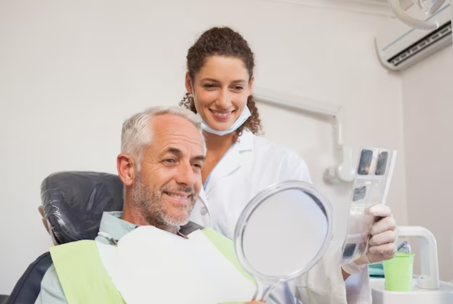 A dentist and a male patient in a dental office, examining teeth in a mirror. The dentist smiles, holding a shade guide.