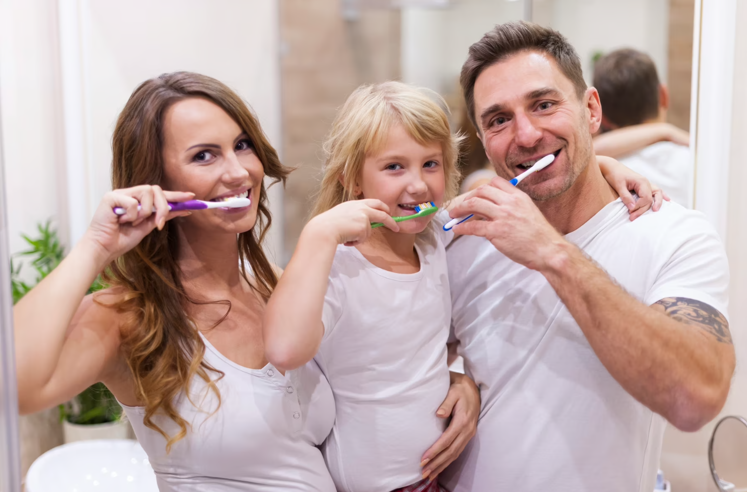 A family of three brushing their teeth in a bathroom; they are smiling, the mother and father flanking the daughter.