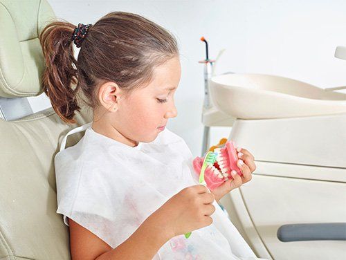 A young girl in a dental chair practices brushing teeth on a model in a dentist's office.