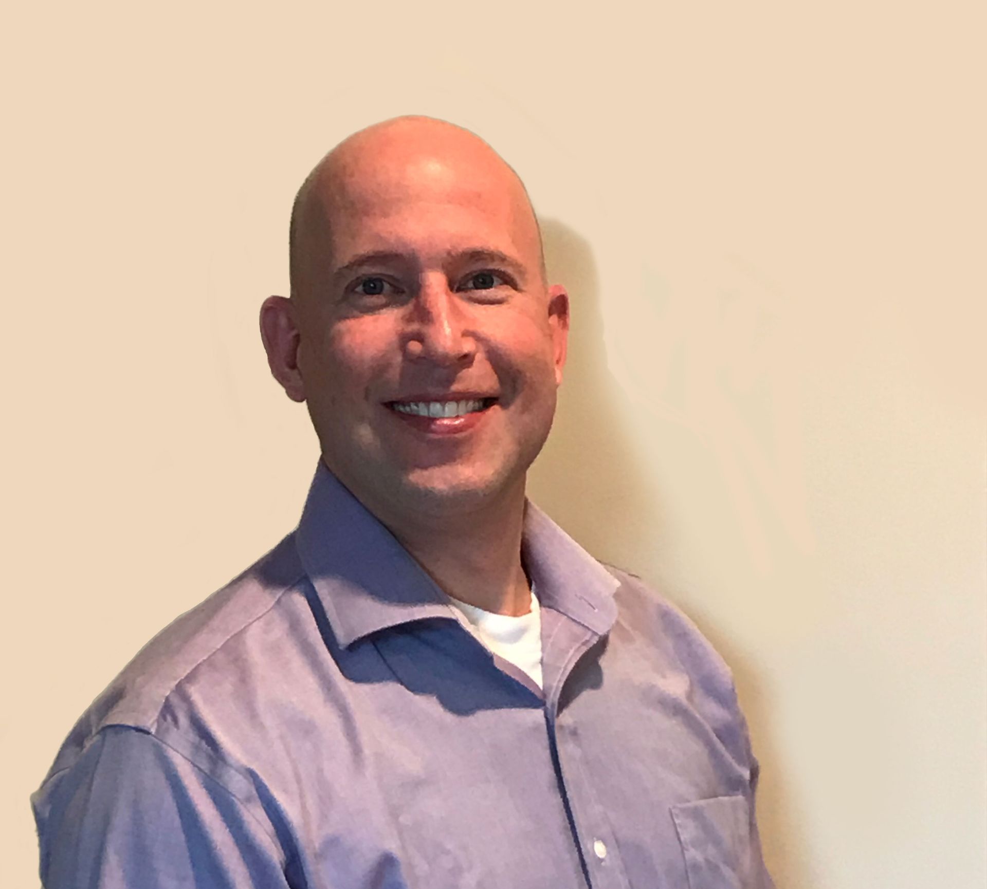 Bald man in a light purple button-down shirt smiles against a beige wall. He wears a white undershirt.