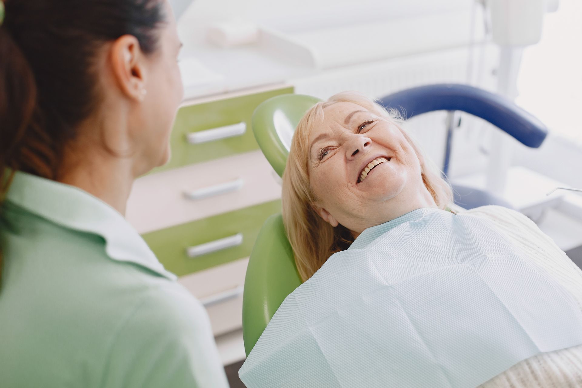 A smiling older woman sits in a dentist chair, wearing a bib. A dental assistant in a green top stands nearby.