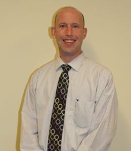 Man in a light-colored shirt and patterned tie smiles at the camera, standing in front of a beige wall.