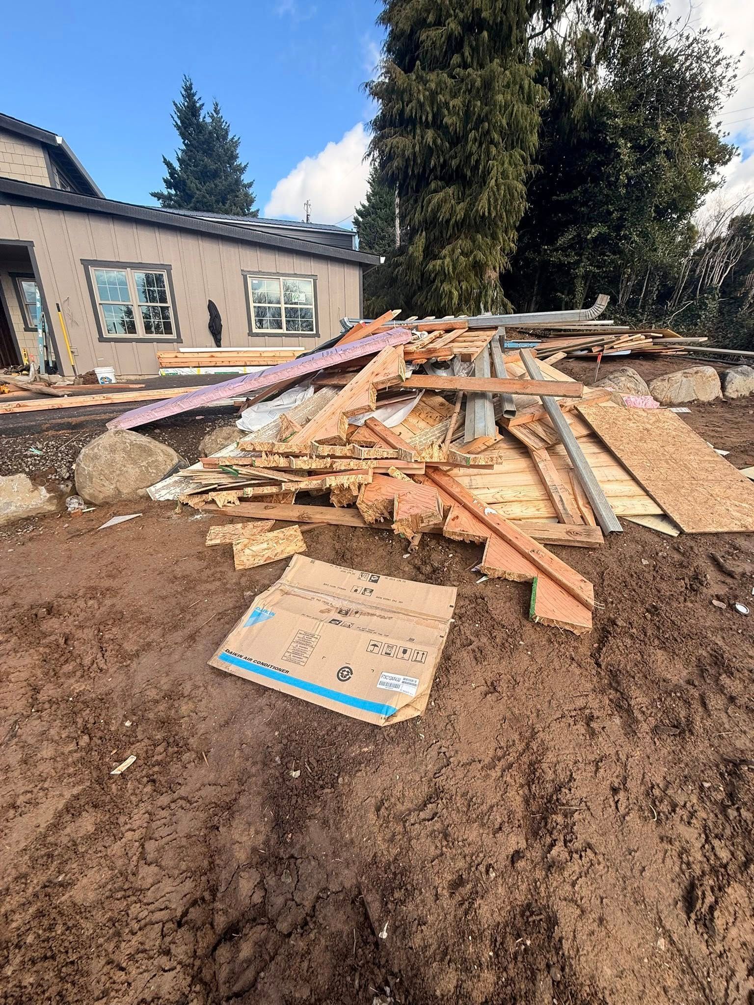 Collapsed wooden debris in a dirt yard beside a house, with trees and blue sky in the background