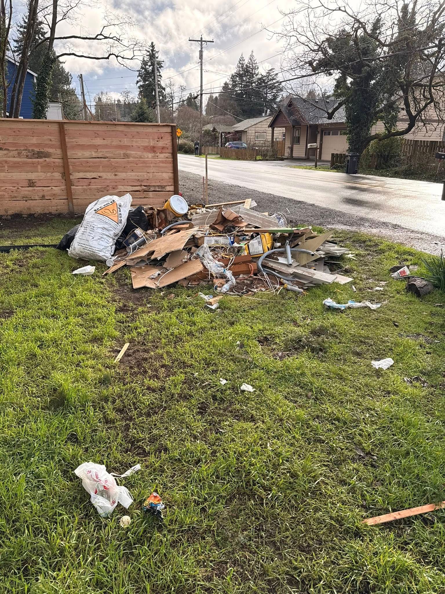 Trash pile on grass beside a road and wooden fence under a cloudy sky