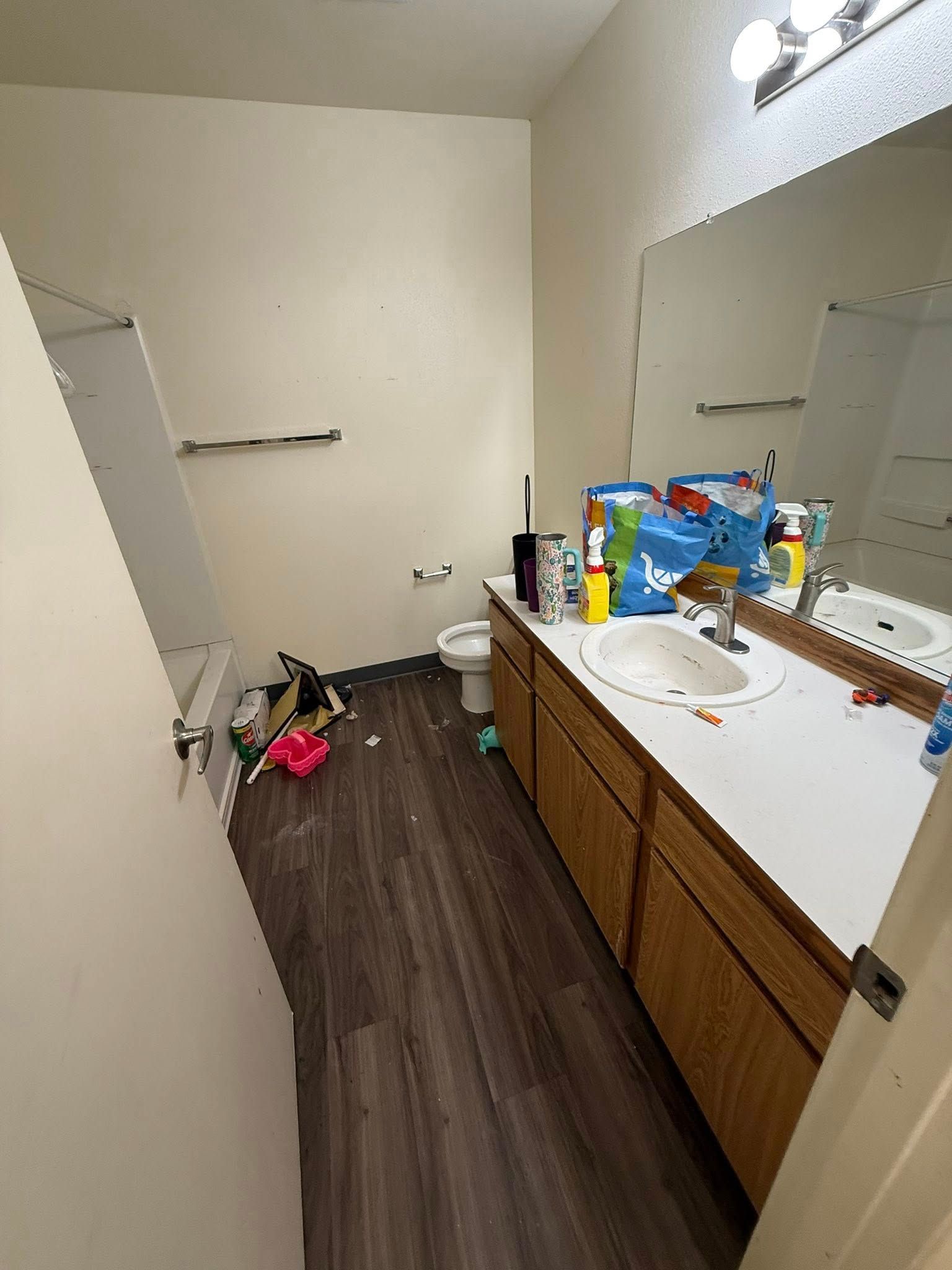 Narrow bathroom with vanity, mirror, toiletries, and a cluttered floor with a pink bag and cleaning items