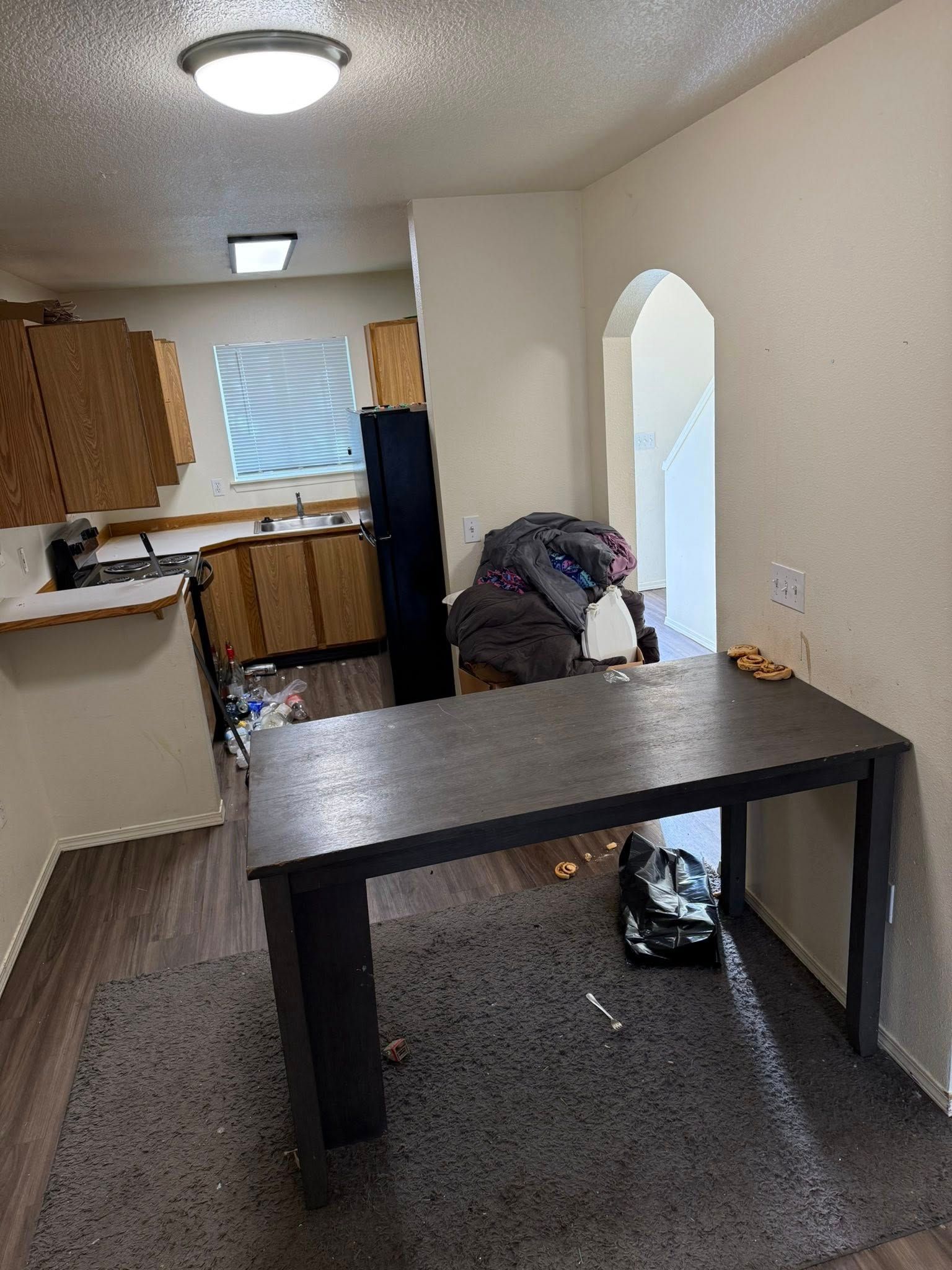 Narrow hallway with a dark table, carpeted floor, kitchen area, and a person bending near the archway.