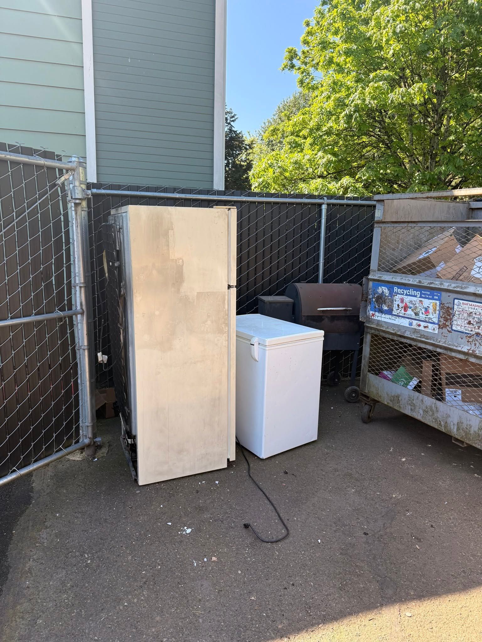 A tall wooden cabinet and white mini fridge sit beside a chain-link fence in an outdoor yard.