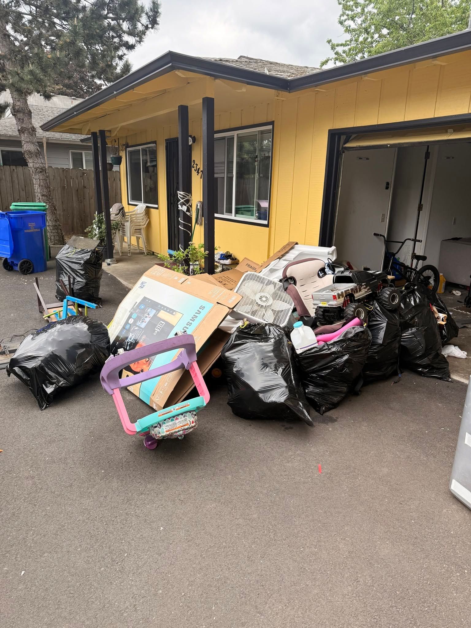 Piles of trash bags and discarded items piled beside a yellow building and open garage.
