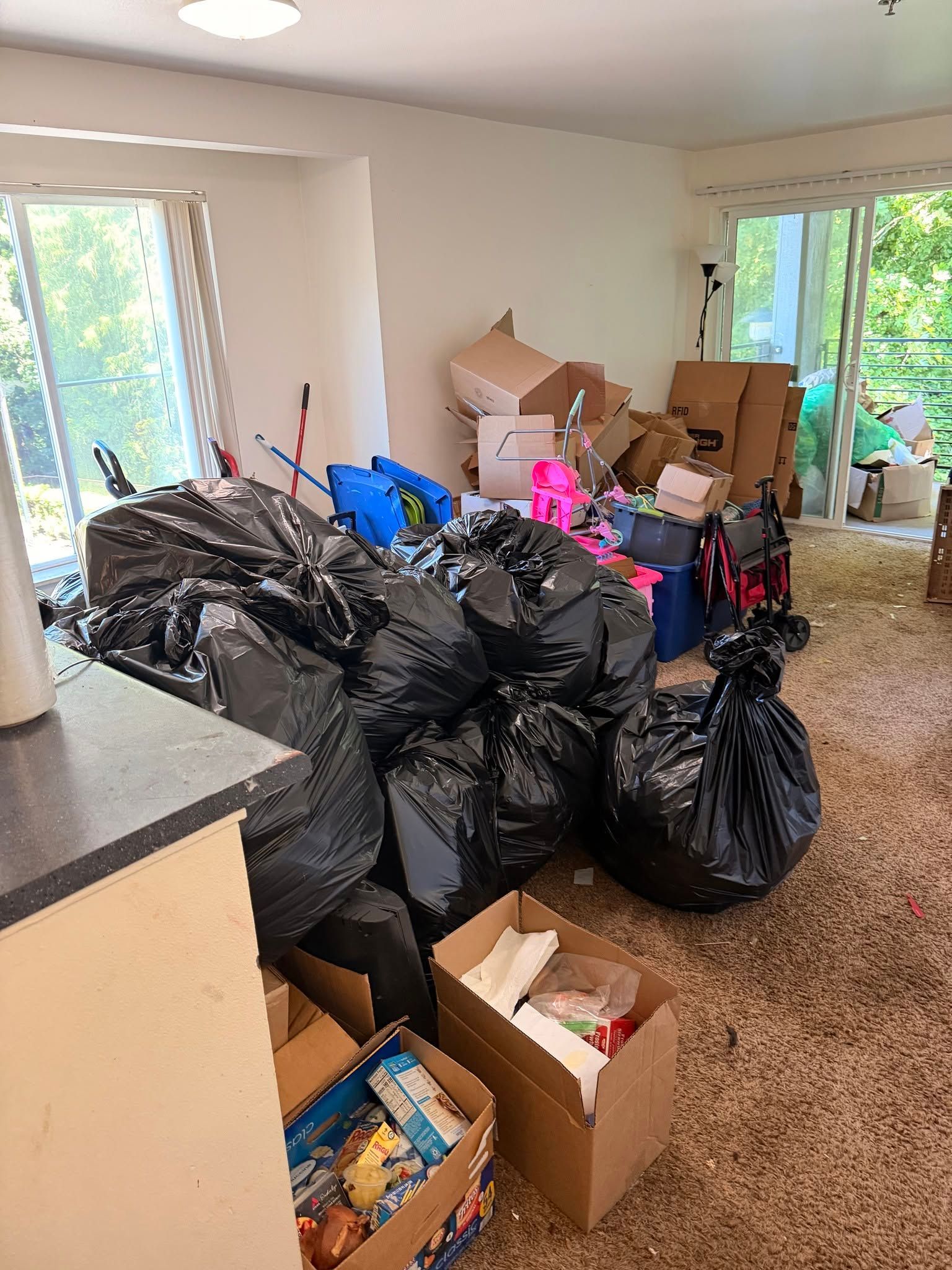 A cluttered room with many black trash bags, cardboard boxes, and household items near large windows.