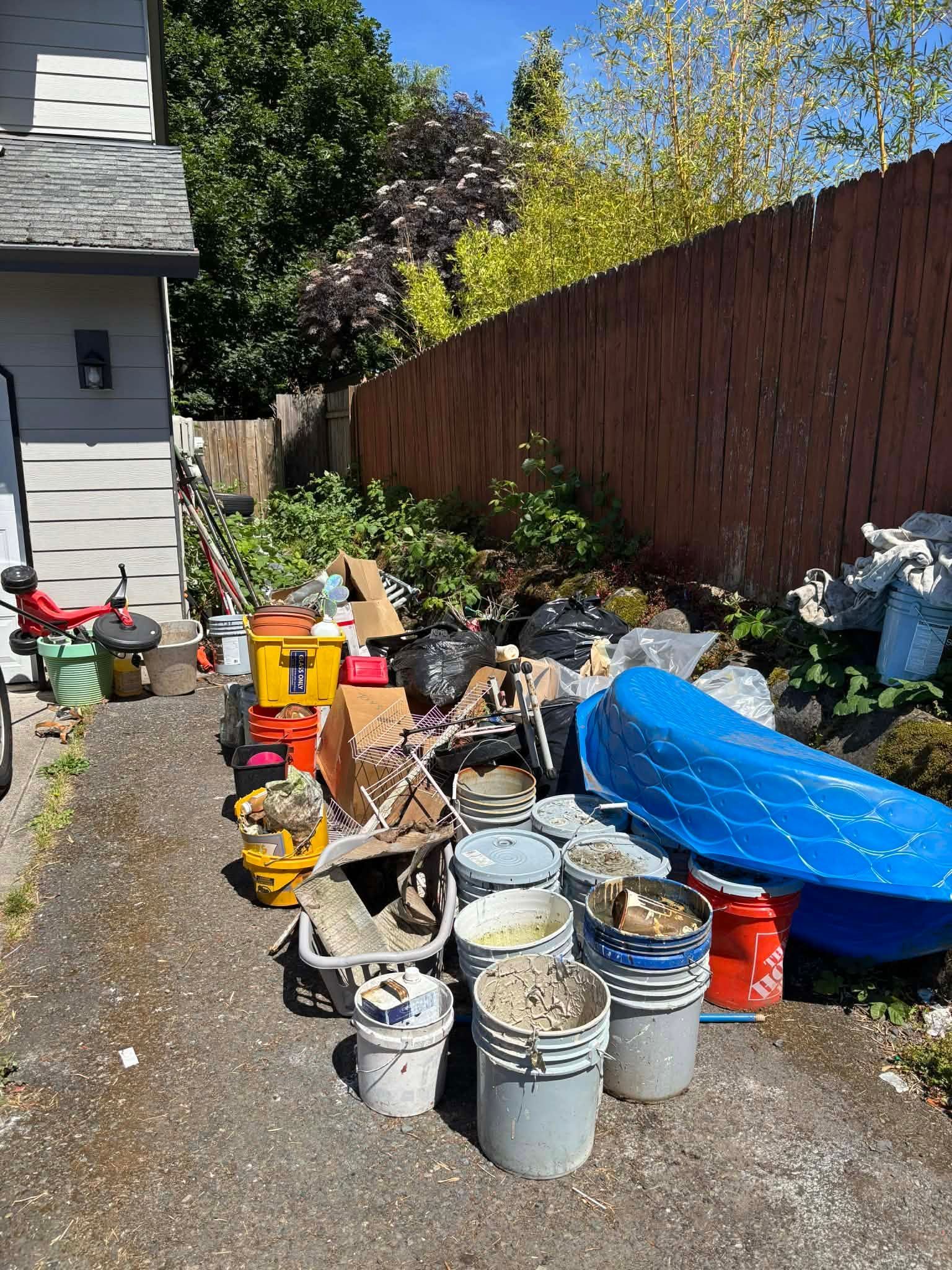 Backyard cluttered with paint cans, buckets, tools, and covered items beside a fence and house.