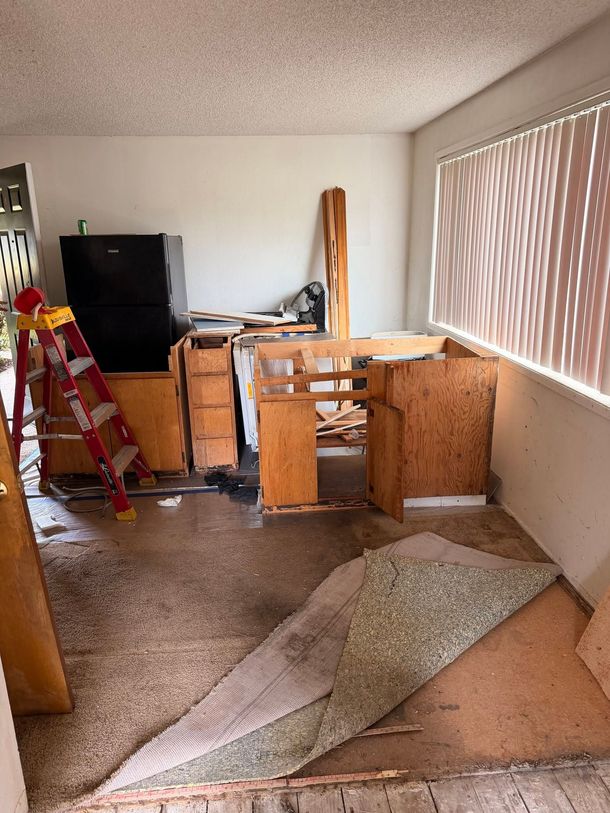 Messy living room with overturned furniture, a ladder, and carpet pieces on the floor near a window with blinds