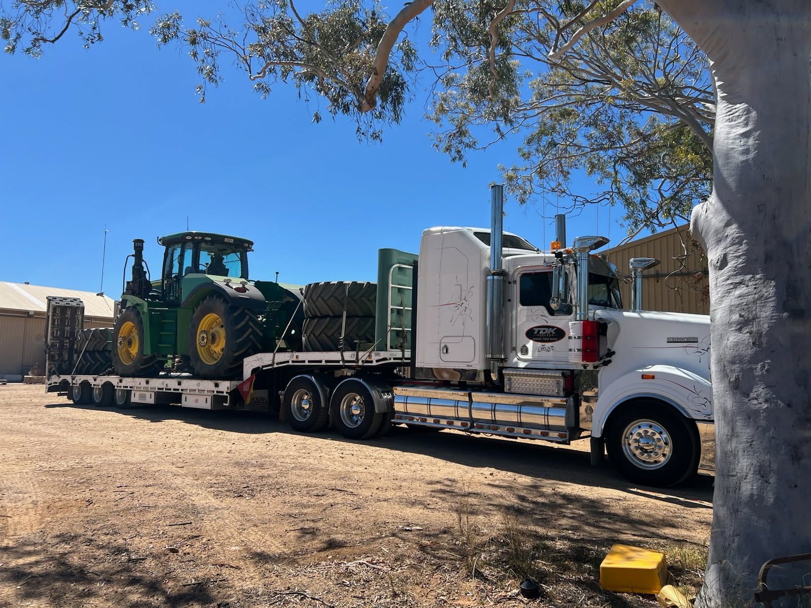White semi-truck hauling a green tractor on a flatbed in a dusty yard under a blue sky