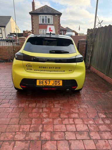 Yellow hatchback car parked on a wet driveway outside a house, rear view with L-plate visible.