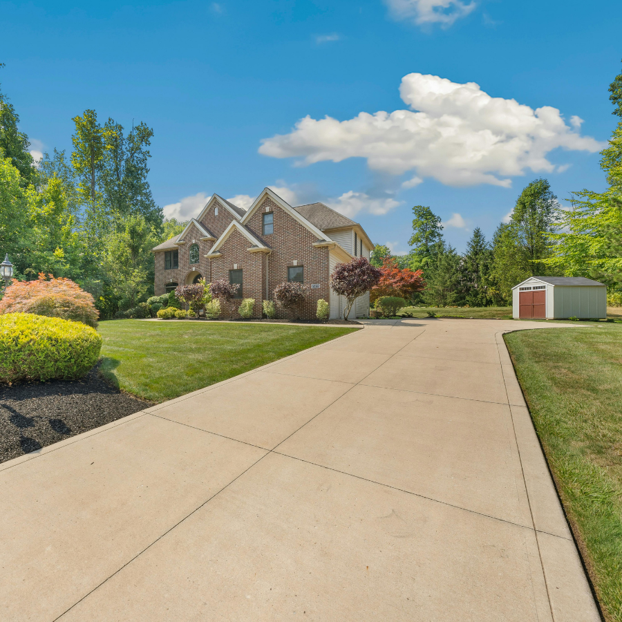 Suburban brick house with a long driveway, green lawn, and blue sky with clouds