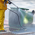 Worker in yellow rain gear pressure-washing a green trash bin on a wet street