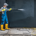 Worker in yellow boots pressure-washes a dark wall with a spray wand.