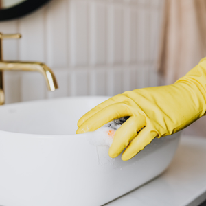 Yellow-gloved hand cleaning a white sink with soap in a bathroom