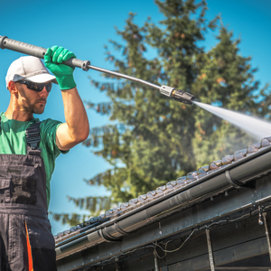 Worker in green gloves pressure-washing a roof under a blue sky