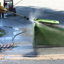 Worker spraying water on a green trash bin beside a street drain, with mist and wet pavement