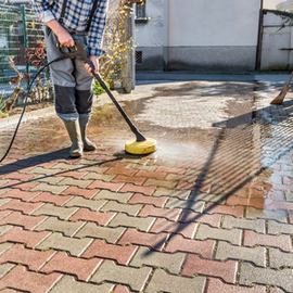 Person using a pressure washer on a sunlit brick driveway next to a house