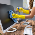 Person cleaning a computer monitor with a blue cloth and spray bottle on a wooden desk