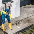 Worker pressure-washing a concrete patio beside a house, wearing blue overalls, yellow boots, gloves, and cap