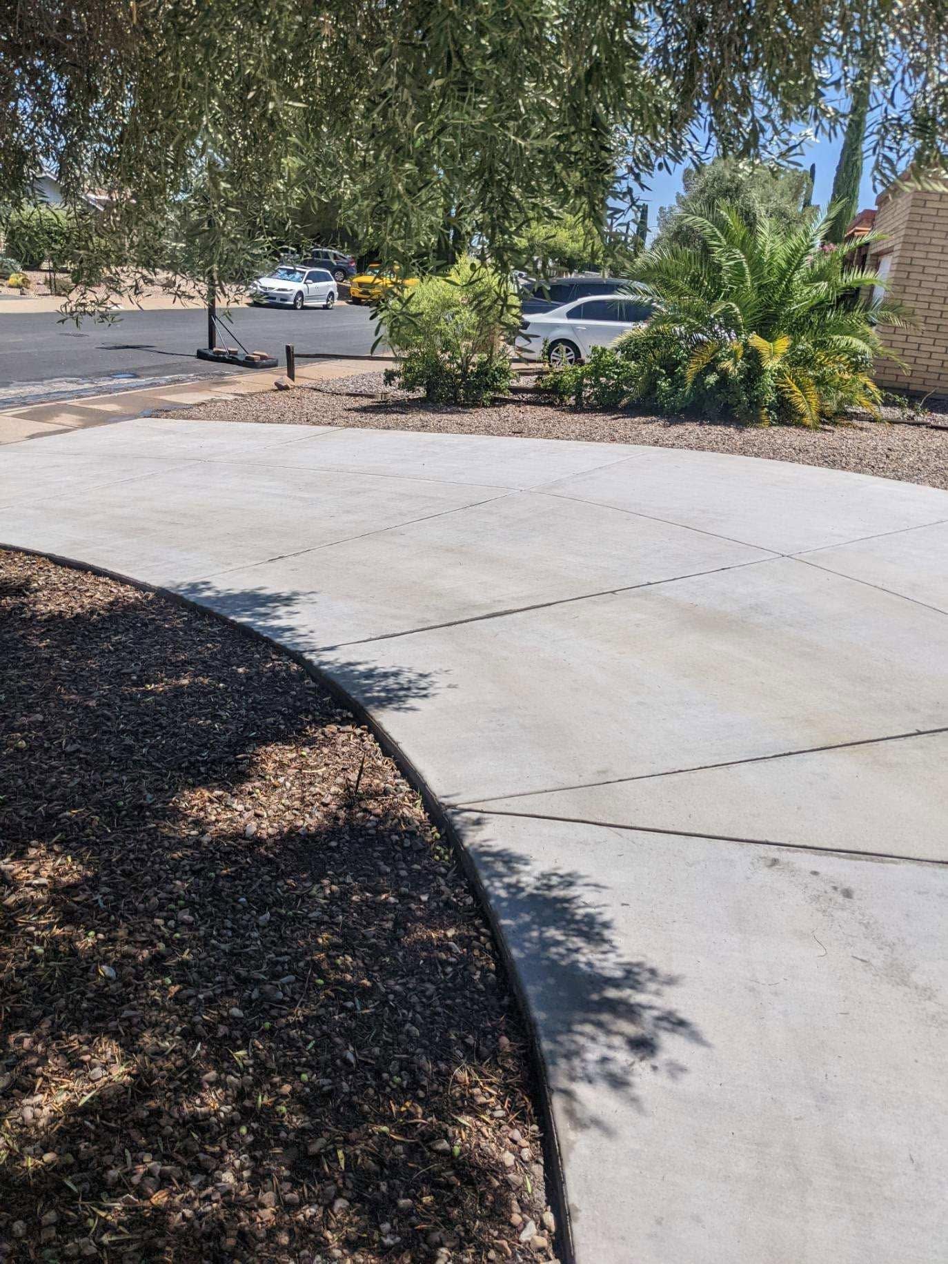 Curved concrete driveway beside a desert landscape with trees and parked cars in the background