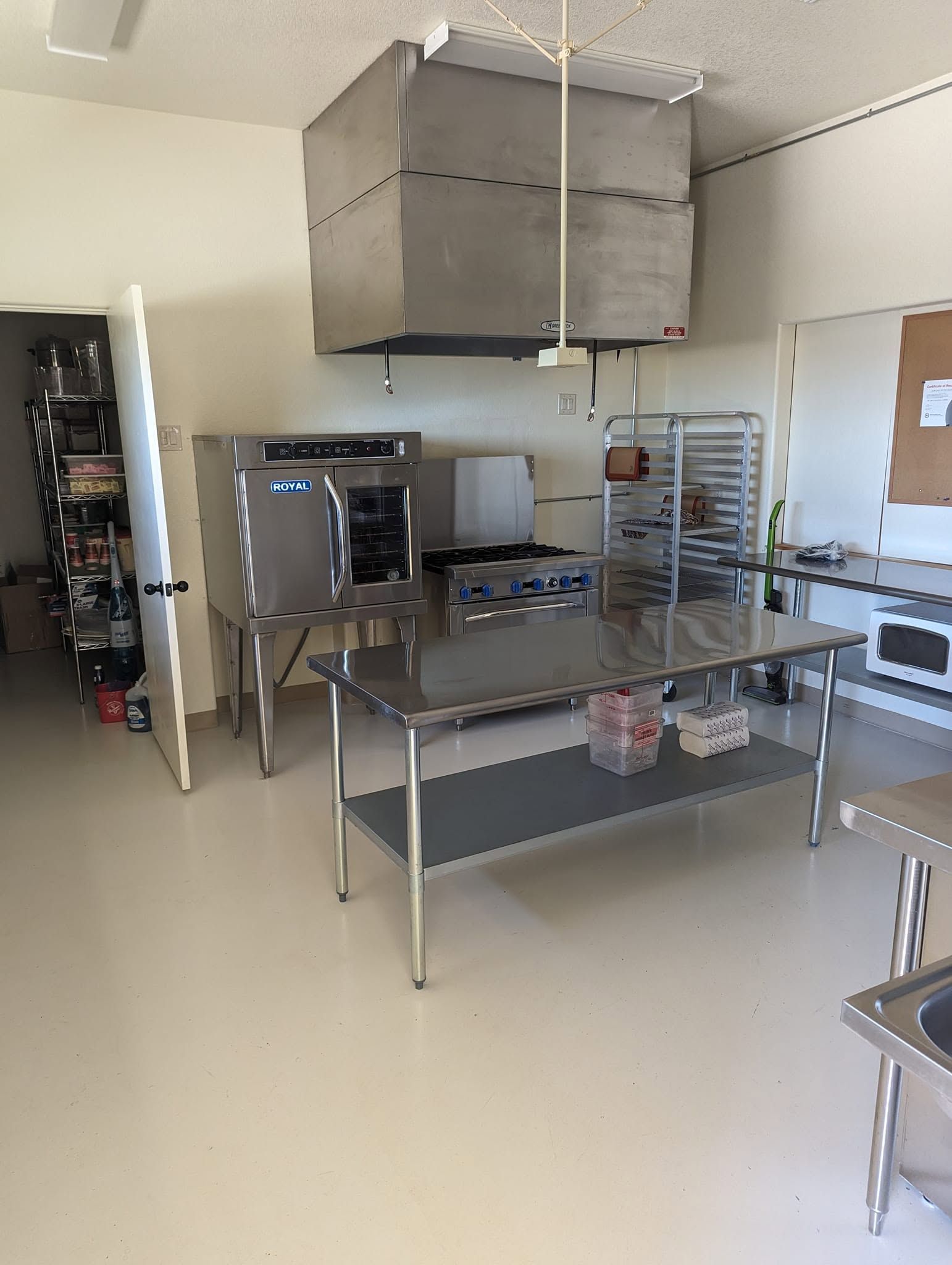Commercial kitchen with stainless steel prep tables, ovens, and a large vent hood on a clean floor.