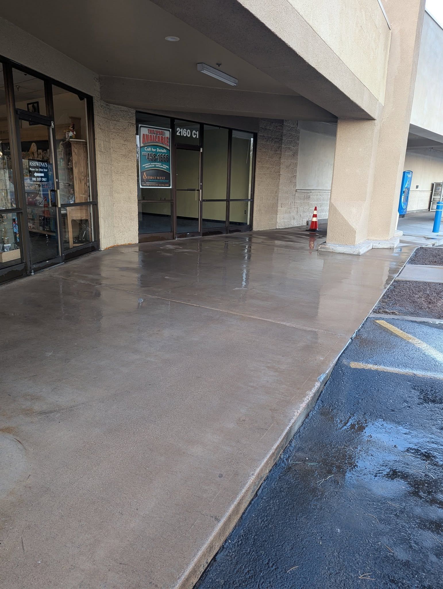 Covered storefront walkway with wet concrete, glass doors, and a red fire extinguisher near the entrance