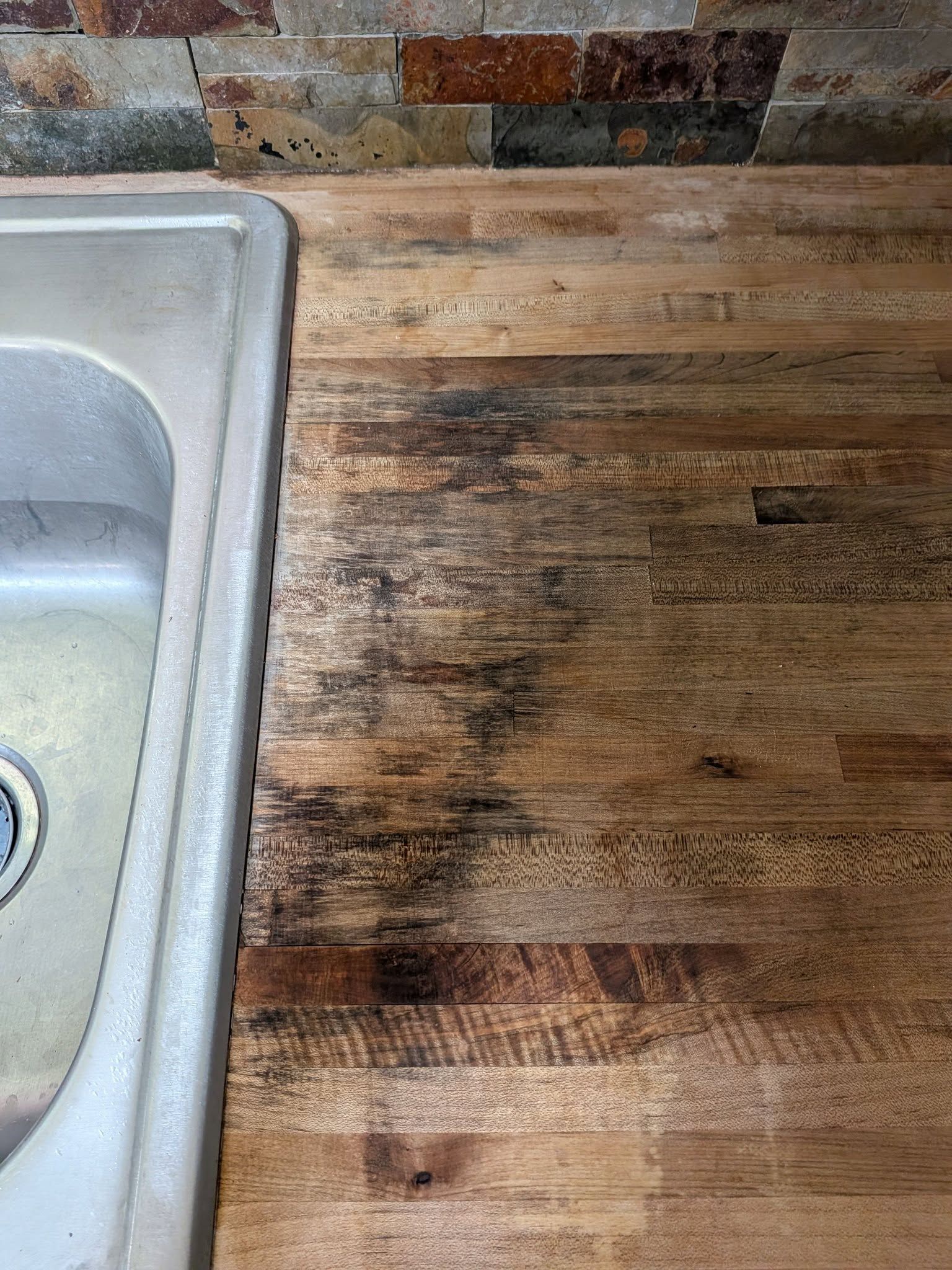 Wooden countertop next to a stainless steel sink, showing visible water stains and wear.