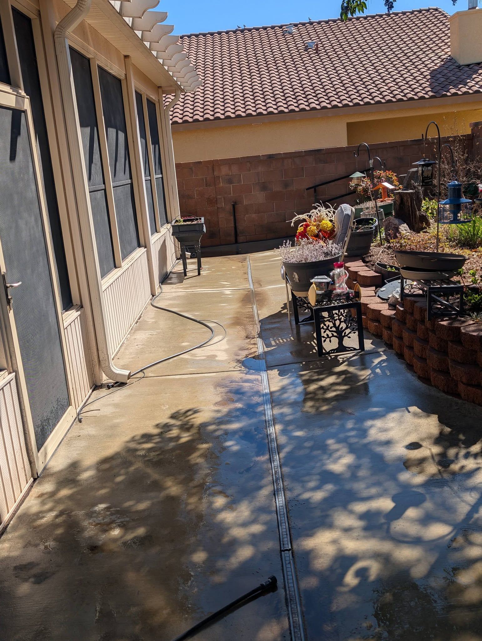 Sunny backyard patio with concrete walkway, sliding glass doors, brick planter, and potted plants beside a house