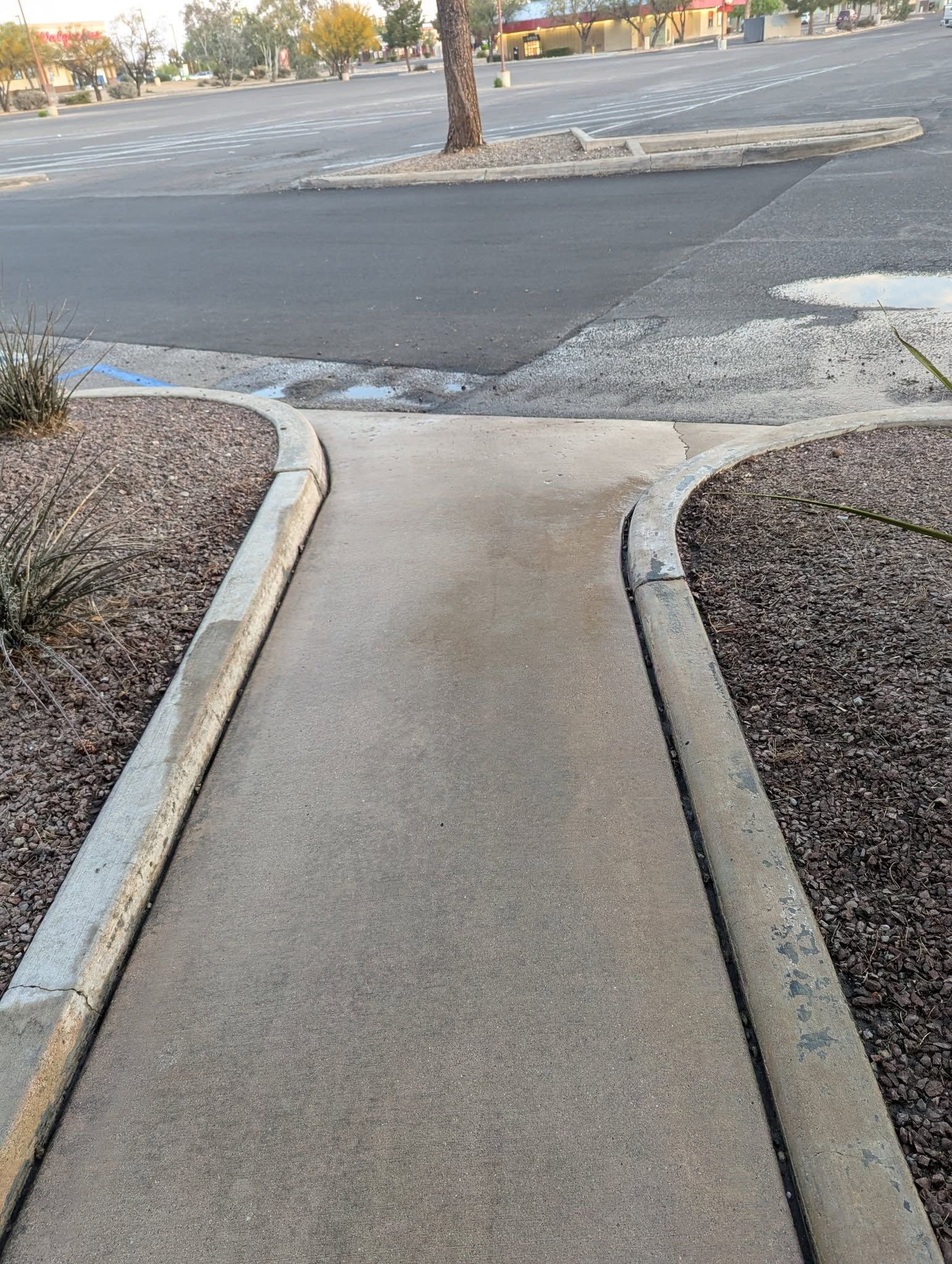 Narrow concrete sidewalk ramp leading to a parking lot curb cut with rough asphalt ahead.