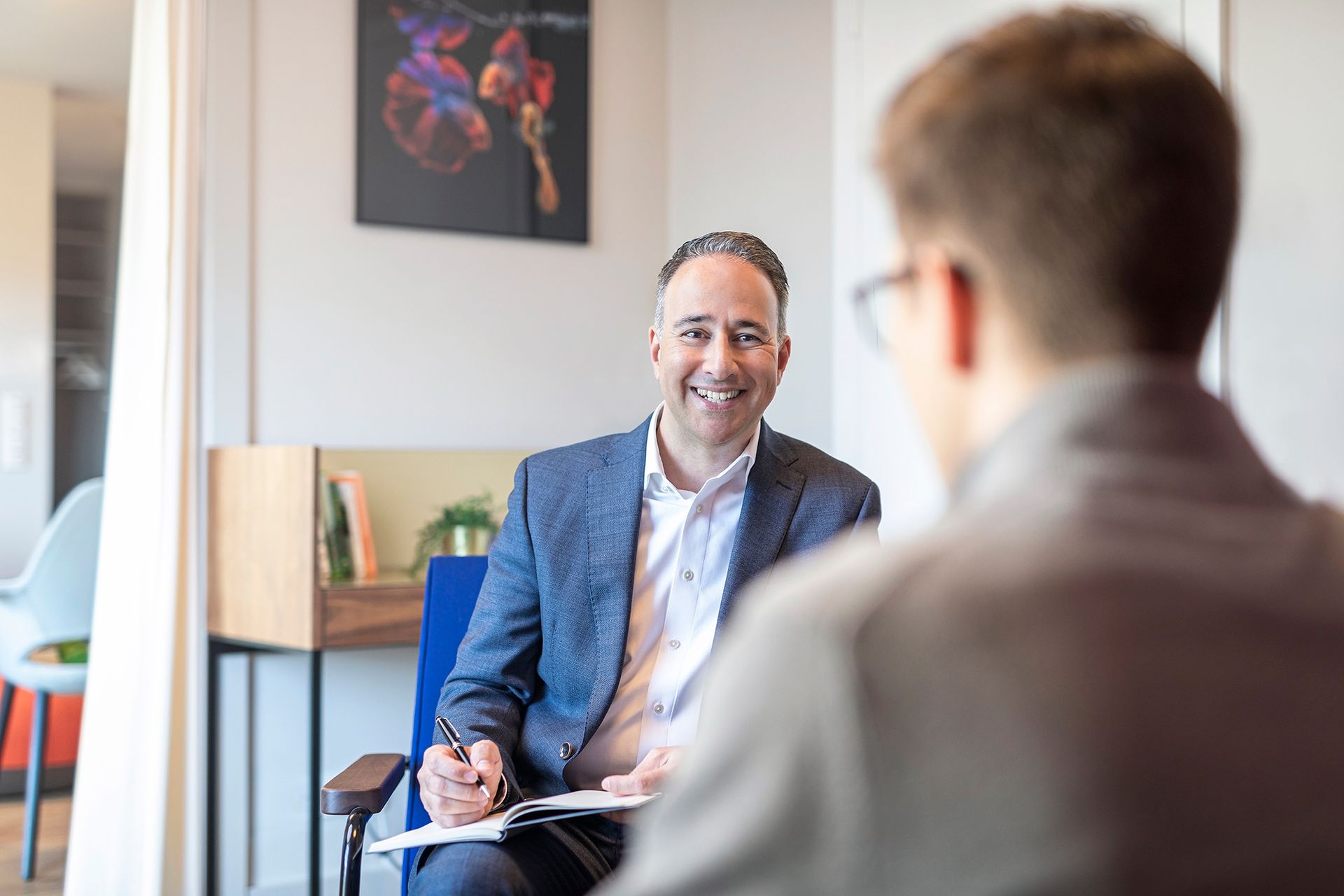 A man in a suit is sitting in a chair talking to another man.