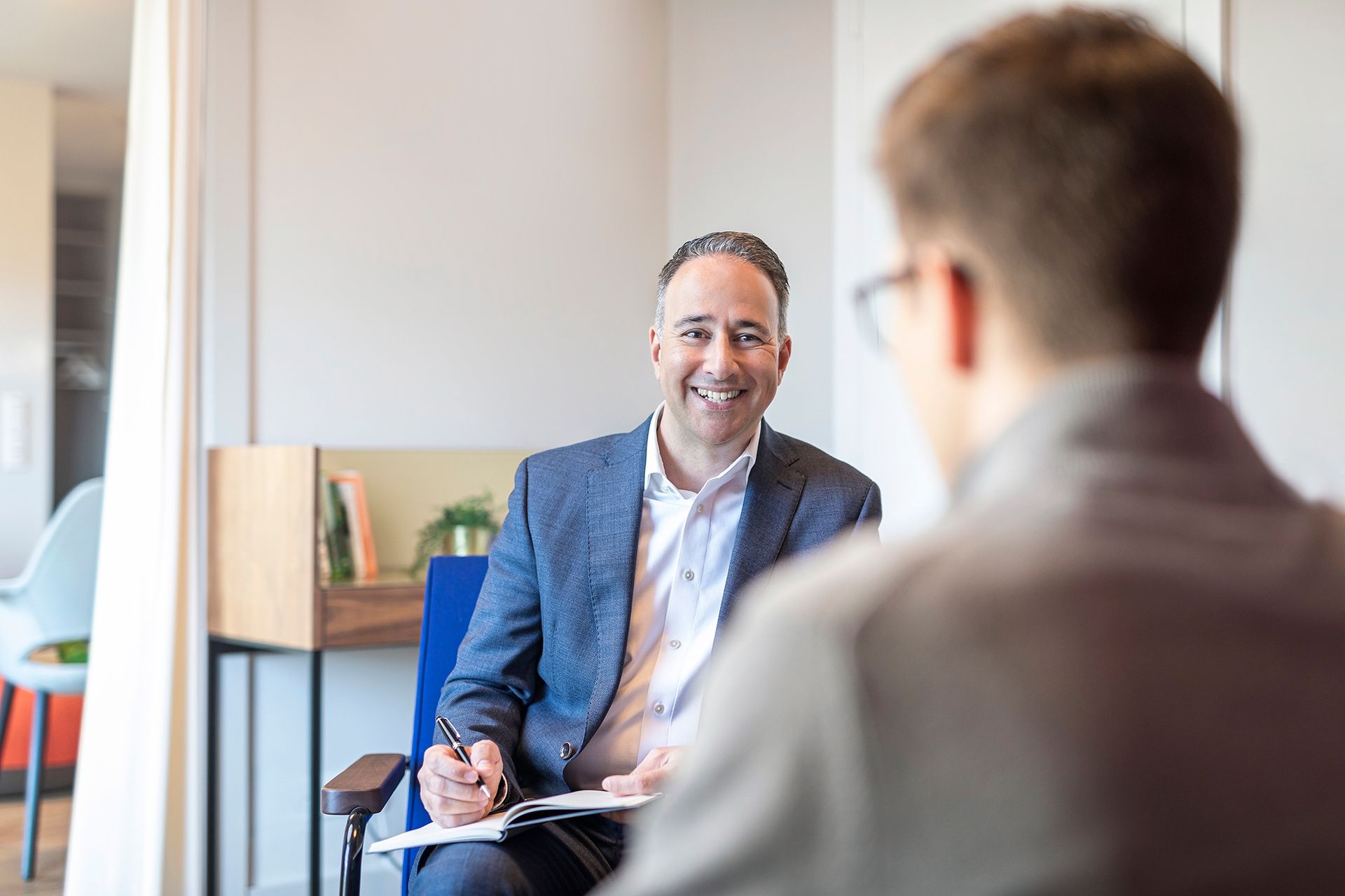 A man in a suit is sitting in a chair talking to another man.
