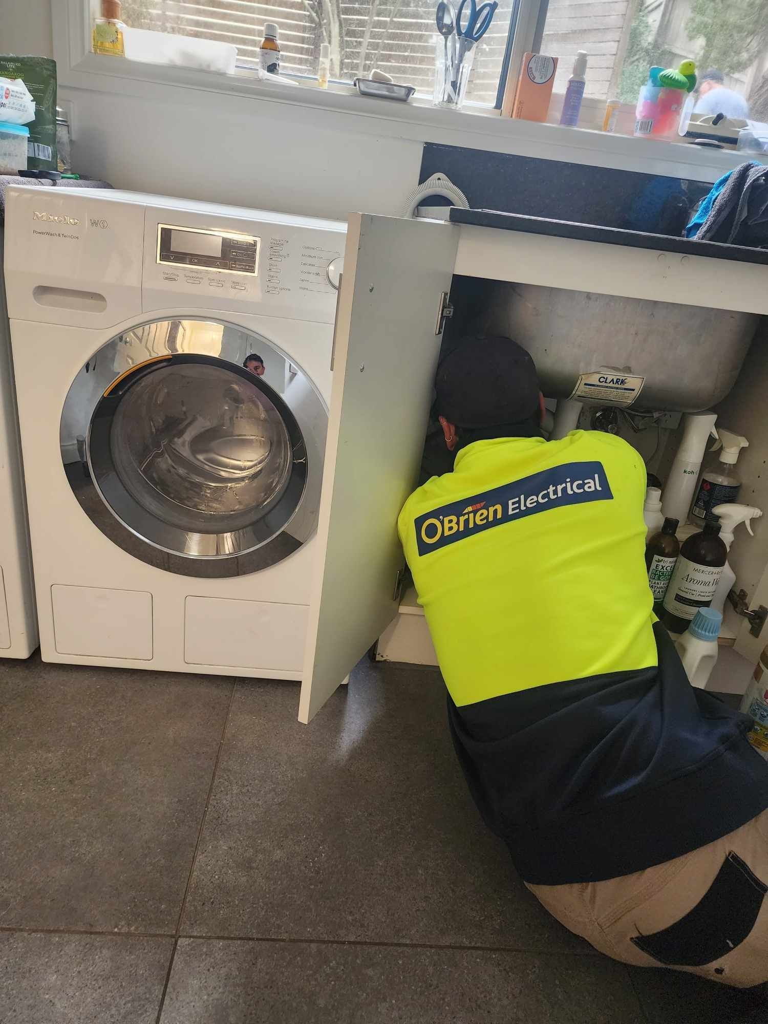 A person in a neon yellow shirt works on plumbing under a sink next to a washing machine.
