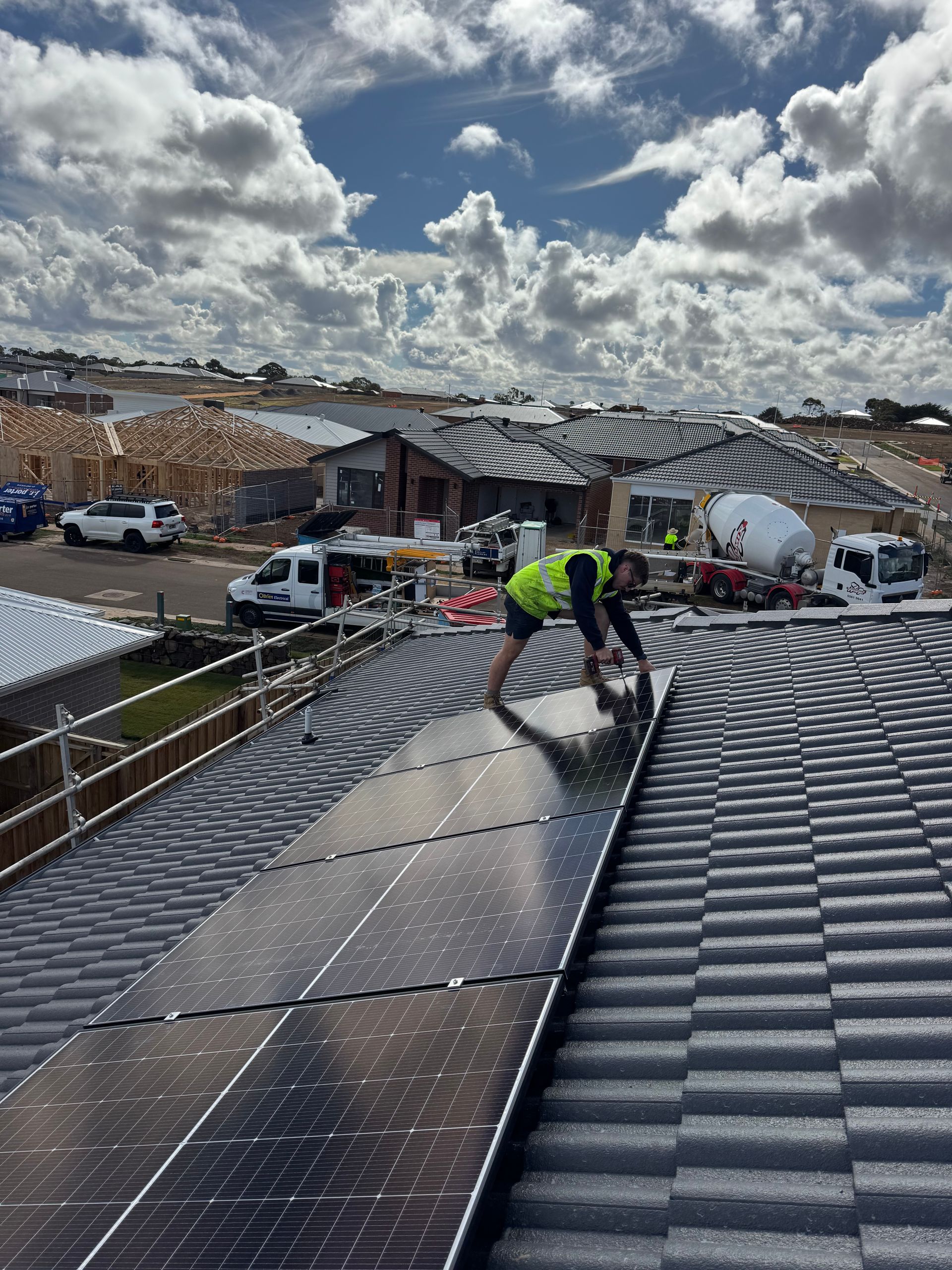 Person installing solar panels on a tiled roof. Construction site in background. Cloudy sky.