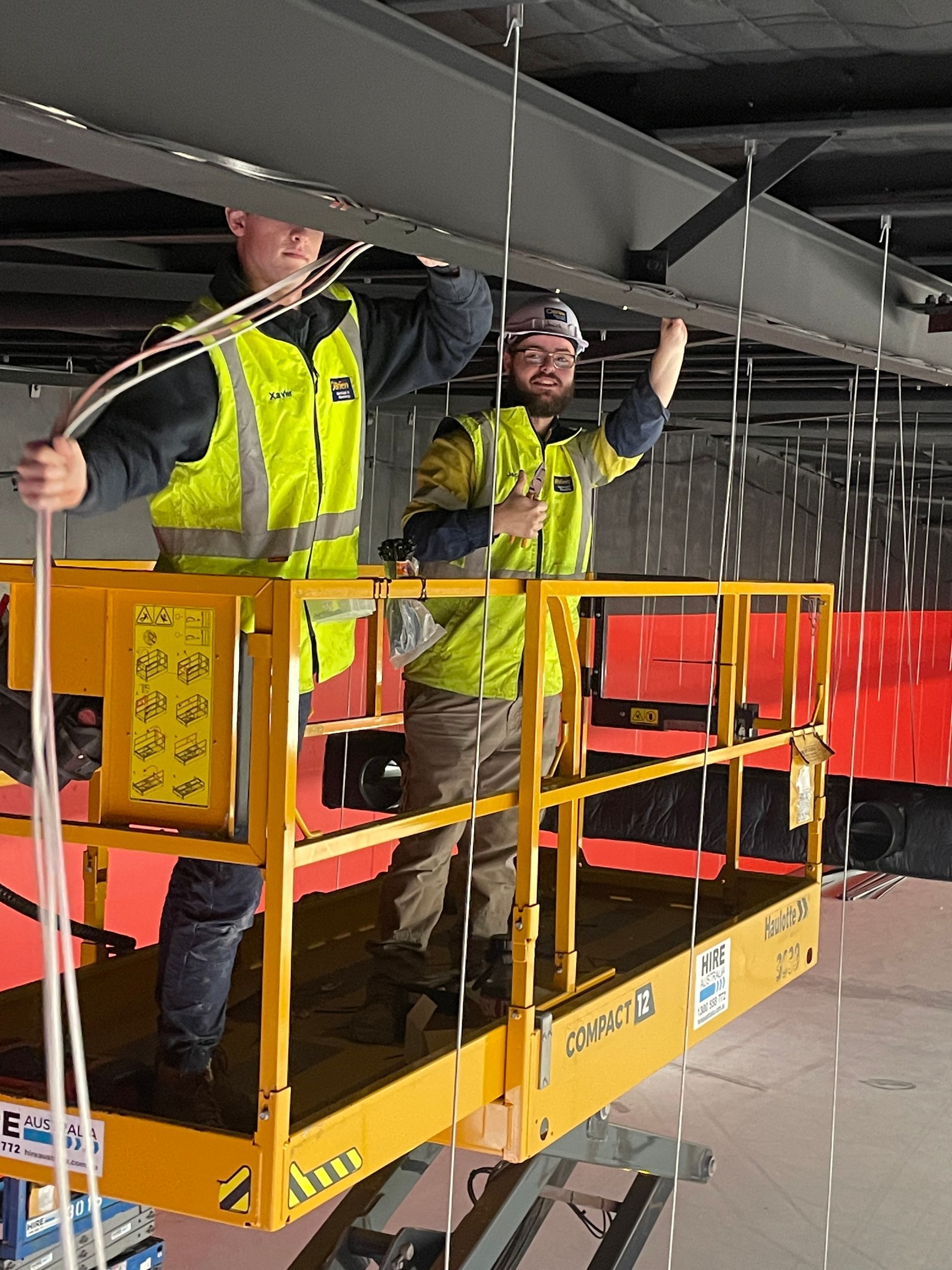 Two workers in safety vests on a lift, installing cables in a construction site.