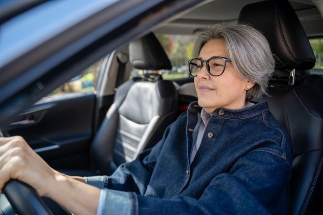 Older person driving a car, wearing glasses and a blue jacket, seen from the front seat