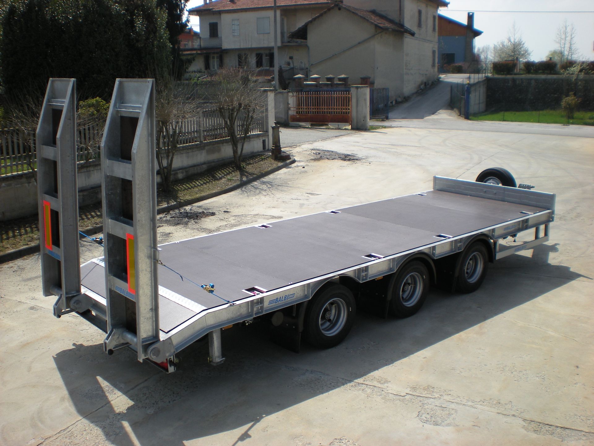 Rimorchio con pianale ribassato, quattro ruote, rampe e piattaforma scura, parcheggiato all'aperto.