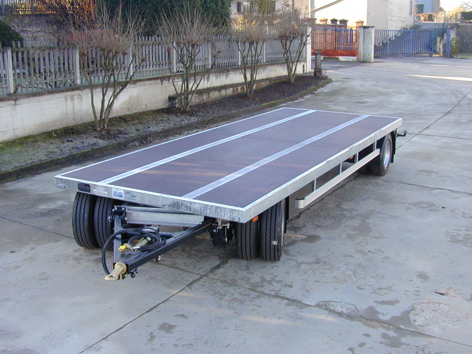 Rimorchio a pianale parcheggiato su una superficie di cemento. Ha un pianale in legno e doppi assi posteriori.