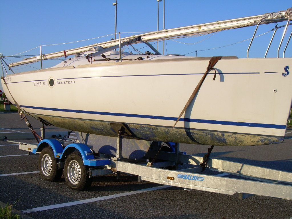 Barca a vela su rimorchio, scafo bianco con striscia blu, parcheggiata sull'asfalto.